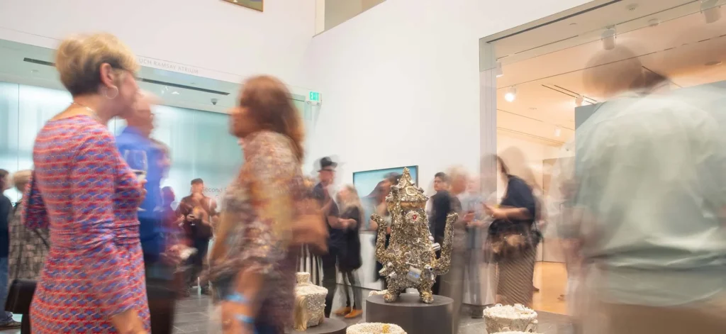 A busy, family friendly art gallery with blurred motion of people walking and talking; sculptural artworks are displayed in the center of the modern, brightly lit room, reminiscent of vibrant Hilliard exhibitions.