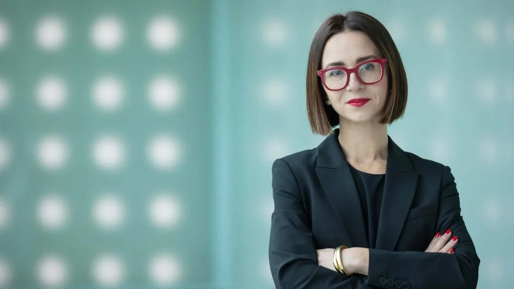 A confident woman with short brown hair, red glasses, and red lipstick stands with arms crossed in a black blazer, against a blurred blue background with white dots—perfectly at home at a Lafayette art museum or other Louisiana museums.