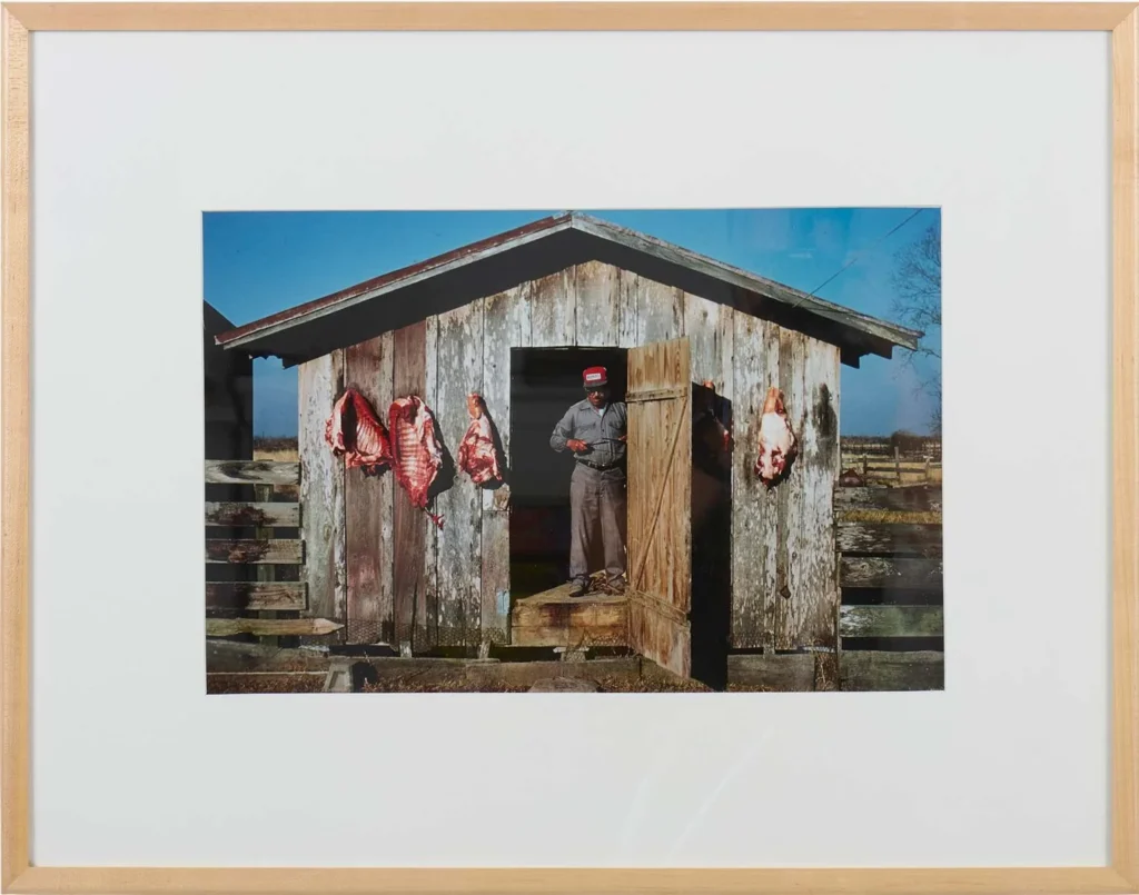 A man in work clothes stands in the doorway of a weathered wooden shed with several large cuts of meat hanging on hooks outside, evoking scenes you might find showcased at Louisiana museums under Hilliard exhibitions. The sky is clear and blue in the background.