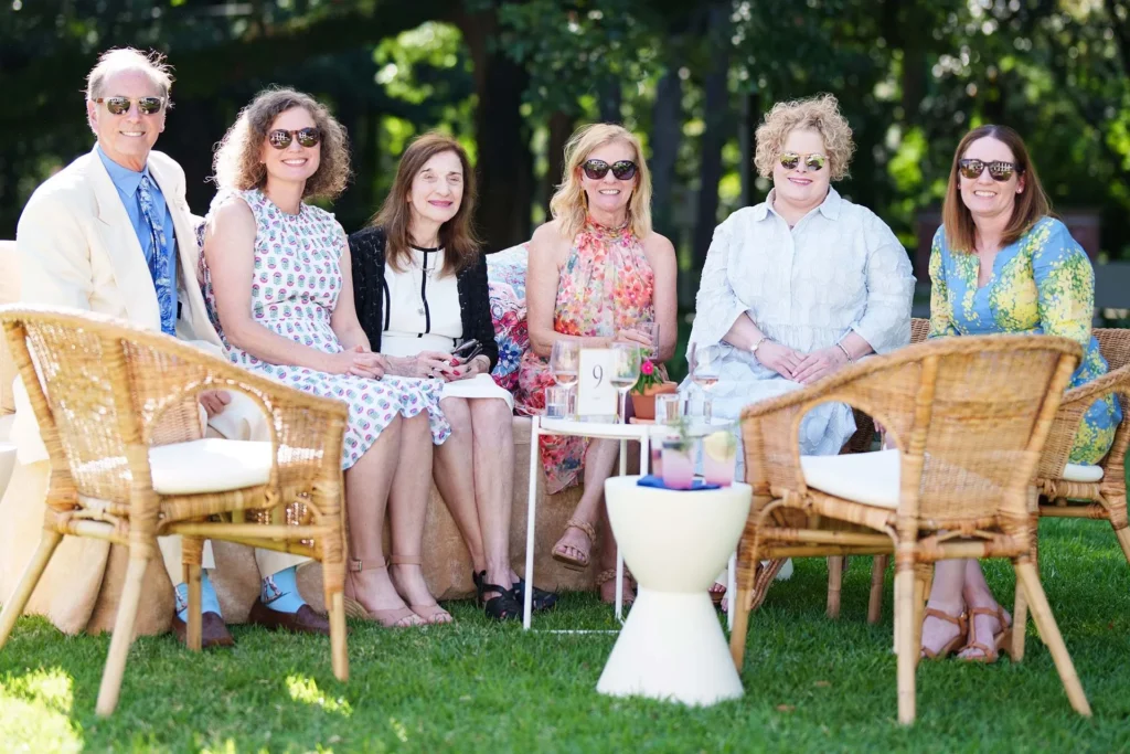 Six adults sit together outdoors on wicker chairs around a small table, smiling at the camera. They are dressed in summer clothing and sunglasses, with green trees and sunlight in the background.