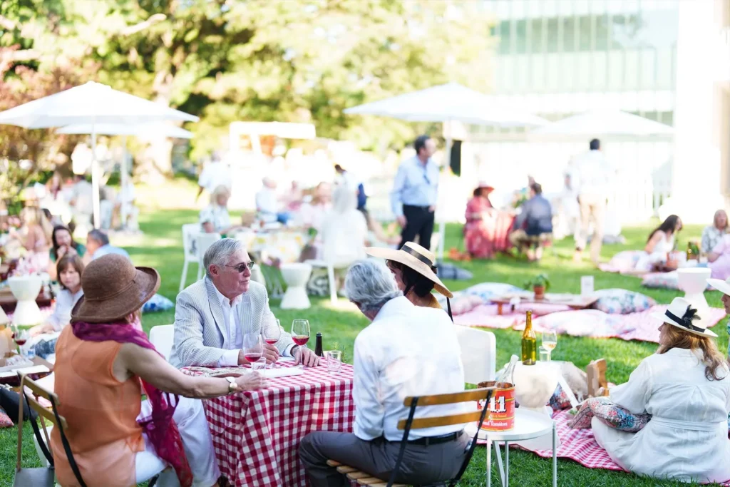 People sit at tables and on picnic blankets on a grassy lawn for an outdoor picnic event. Some wear hats and sunglasses, and food and drinks are on the tables. White umbrellas provide shade in the background.