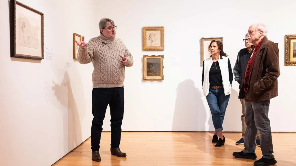 A man in a beige sweater gestures while speaking to three people in the Hilliard Art Museum. They stand on a wooden floor, surrounded by framed artwork hanging on white walls at this vibrant Lafayette art museum.