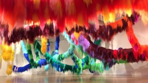 Colorful, fuzzy, yarn-like strands in red, orange, yellow, green, blue, and purple hang from a ceiling and twist across the gallery at the Hilliard exhibitions, creating a vibrant, immersive art installation.