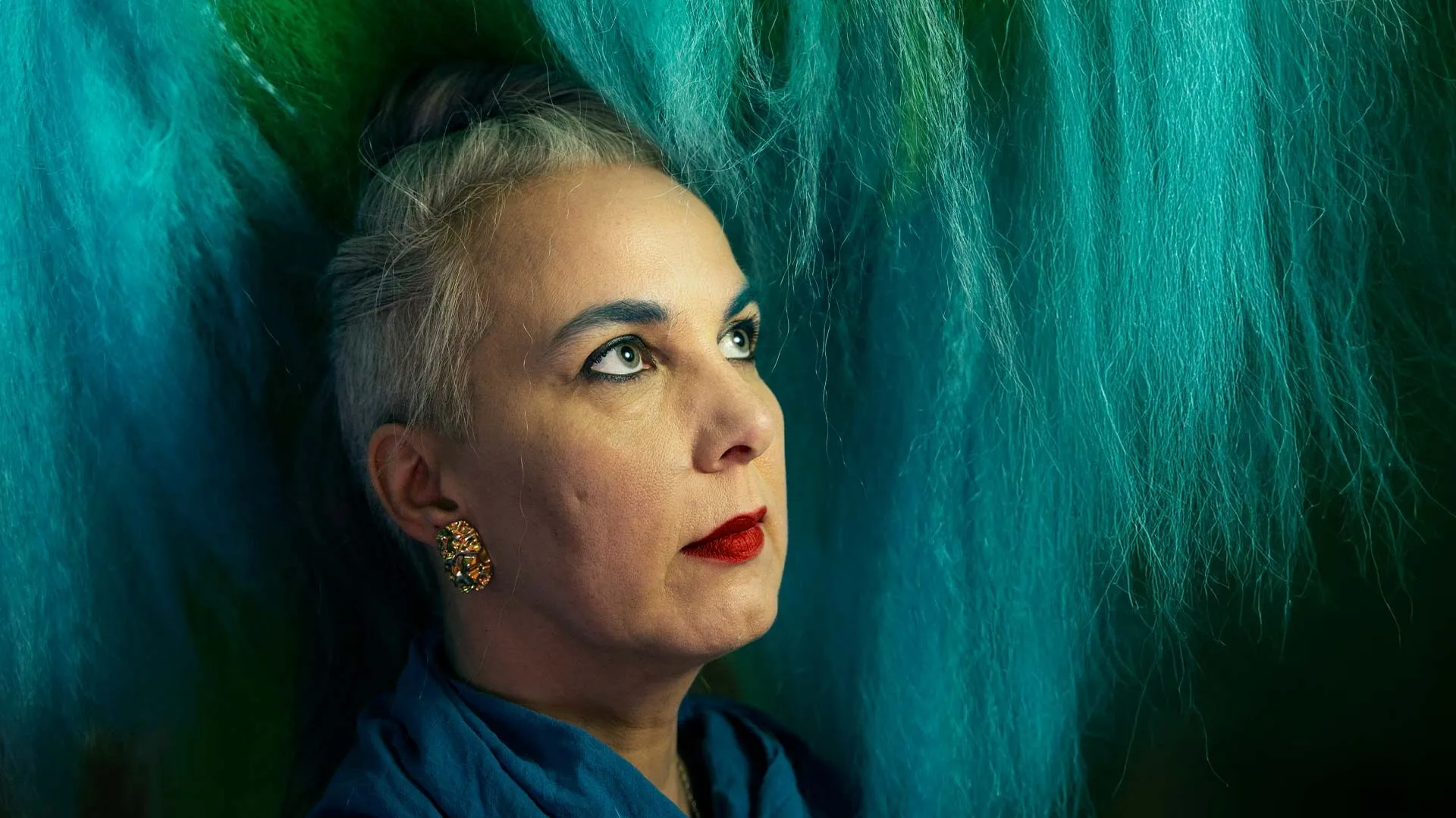 A woman with gray hair, blue eyebrows, and red lipstick looks upward. She wears a blue scarf and gold earrings, surrounded by vibrant blue-green fibers—echoing the creative spirit found in local art Lafayette offers.