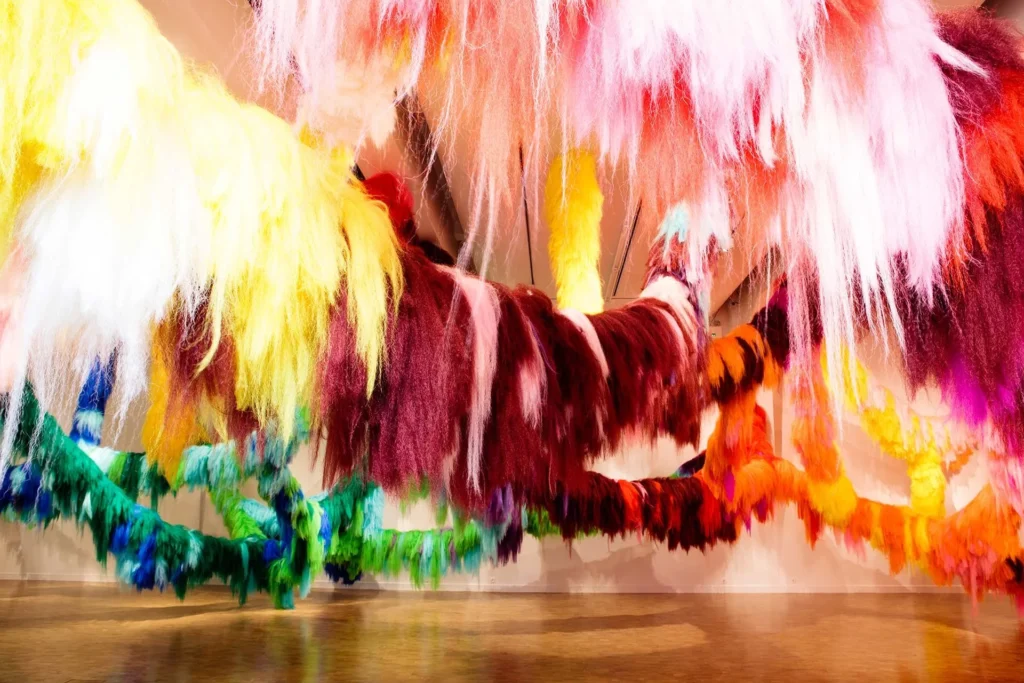 Colorful, fluffy, yarn-like sculptures in red, pink, yellow, green, and orange hang from the ceiling at the Lafayette art museum, creating an immersive installation with a reflective floor that’s perfect for a family friendly outing.