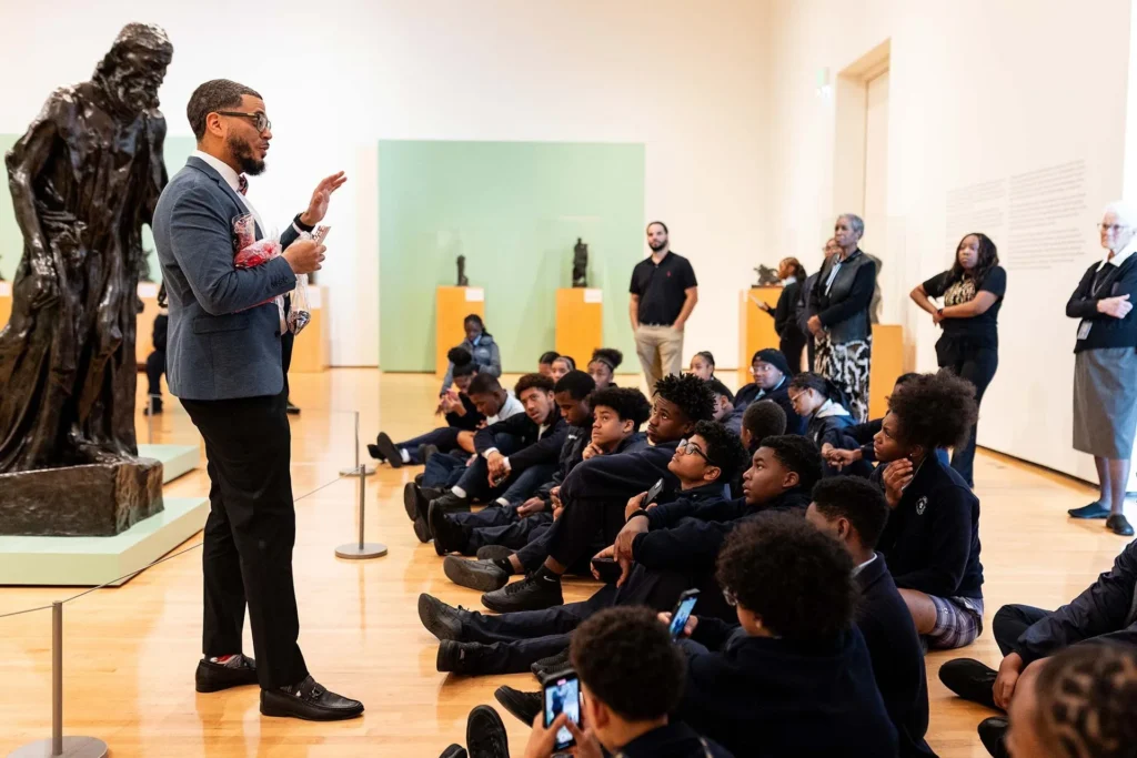 A man speaks to a group of seated students in a museum gallery. Several adults stand nearby, and a large bronze sculpture is displayed next to the speaker. The students appear attentive and engaged.