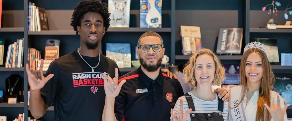 Four people stand together smiling in front of a bookshelf at the Hilliard Art Museum. Two hold up a hand sign, one wears a "Ragin’ Cajuns Basketball" shirt, and another wears a crown and sash, creating a Lafayette family friendly moment.