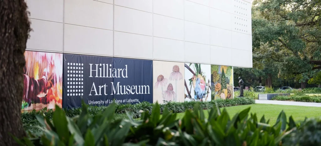 A large sign on the exterior wall of a building reads Hilliard Art Museum, University of Louisiana at Lafayette, with colorful artwork panels displayed next to it, surrounded by green plants and trees.