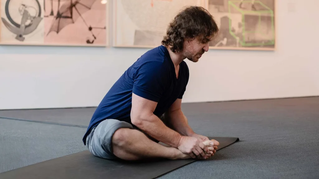 A person with medium-length hair sits on a yoga mat indoors, stretching forward and holding their feet, with artworks from local art Lafayette and Hilliard Art Museum exhibitions visible on the wall behind them.