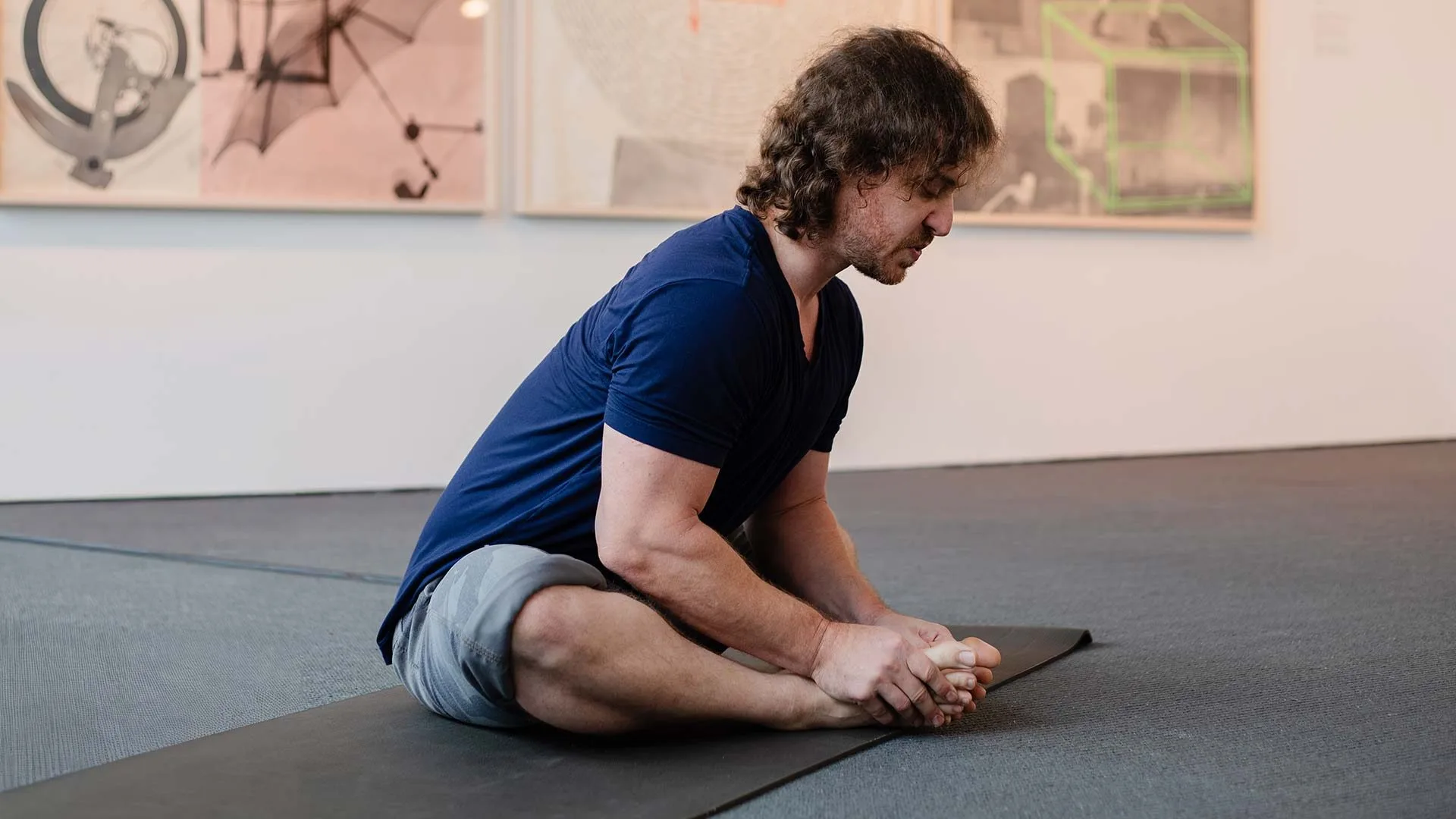 A person with medium-length hair sits on a yoga mat indoors, stretching forward and holding their feet, with artworks from local art Lafayette and Hilliard Art Museum exhibitions visible on the wall behind them.