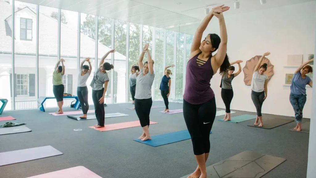 A group of people participate in a yoga class indoors, standing on mats and stretching with arms raised, in a bright room with large windows and natural light.