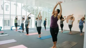 A group of people participate in a yoga class indoors, standing on mats and stretching with arms raised, in a bright room with large windows and natural light.