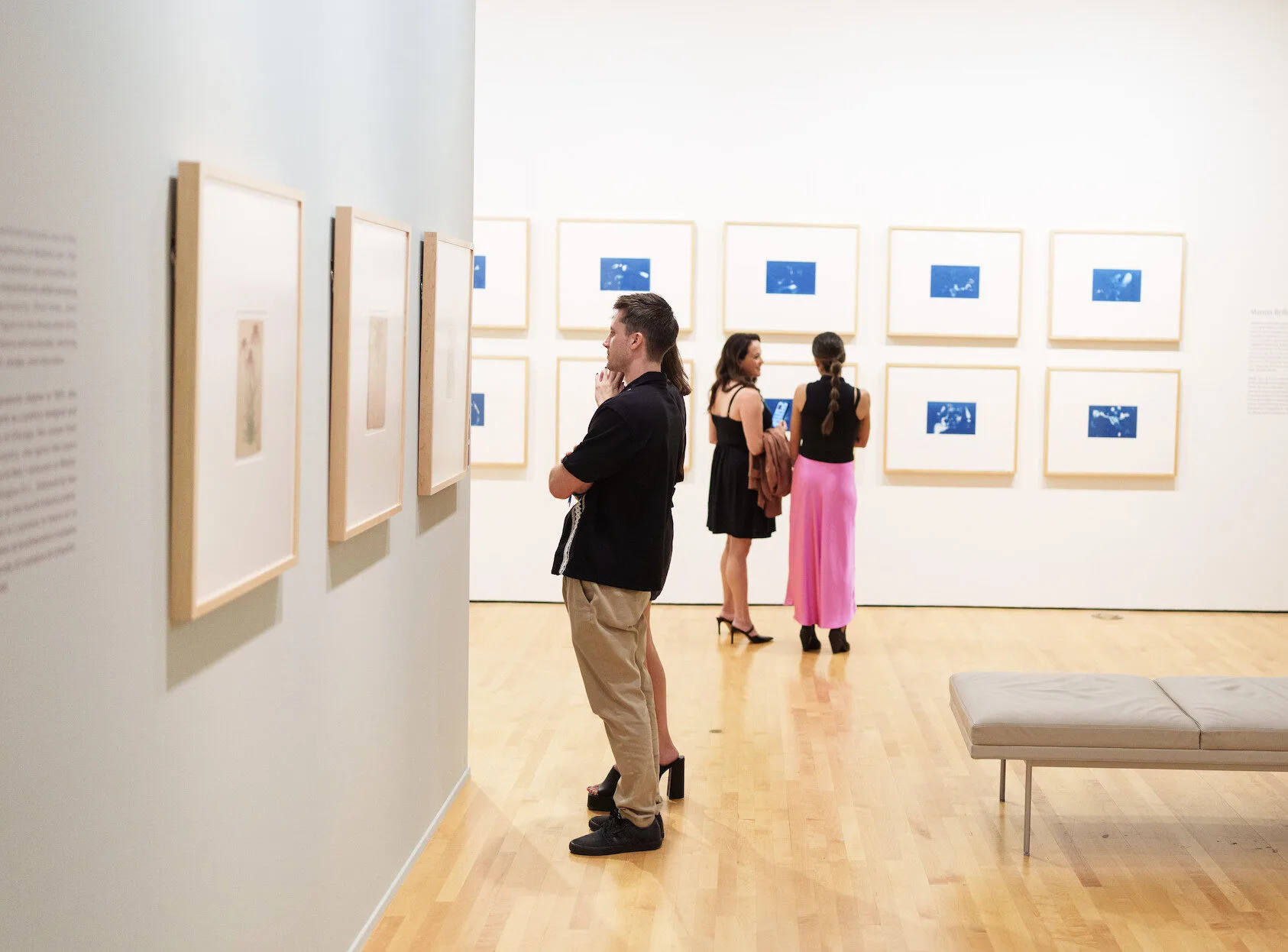 People view framed artwork on white walls in a modern art gallery. Three women and one man stand and observe the pieces closely. The gallery has a light wooden floor and minimal seating.