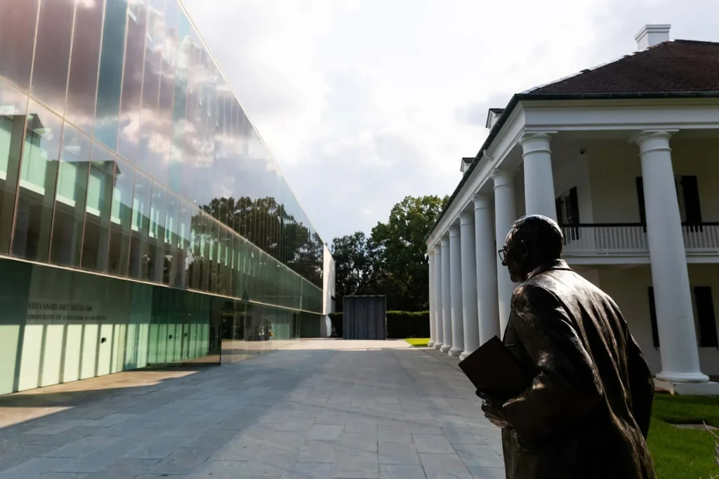 A bronze statue of a person holding a book stands in front of a modern glass building and a white-columned historic building, connected by a stone walkway under a partly cloudy sky.