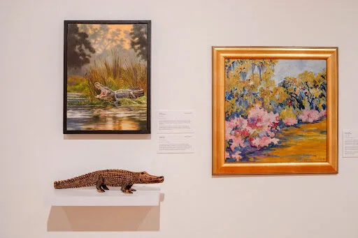 Two framed artworks hang on a wall: one depicts alligators in a swamp, the other is a colorful landscape with trees and flowers. Below them, a textured alligator sculpture rests on a white shelf.