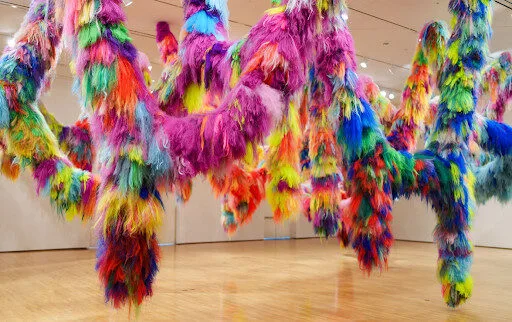 Colorful, furry, vine-like sculptures hang from the ceiling in an art gallery, featuring vibrant shades of pink, blue, yellow, orange, green, and purple, set against white walls and a shiny wooden floor.