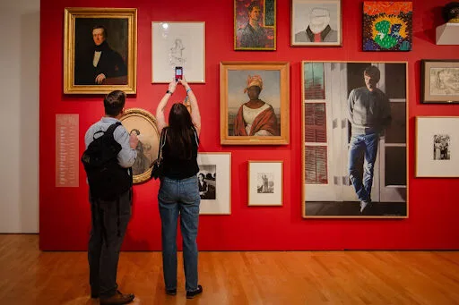 Two people stand in front of a red wall covered with framed artworks; one person holds up a phone to take a picture while the other looks at the art. The floor is wooden, and the art varies in style and size.