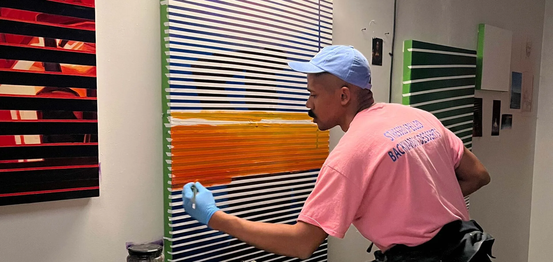 A person wearing a pink shirt, blue cap, and blue gloves paints orange and white horizontal stripes on a canvas with black lines, displayed on a wall alongside other artwork.