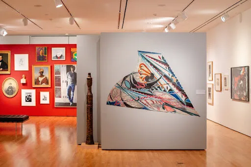 A contemporary art gallery features colorful abstract and portrait paintings on red and white walls, wooden sculptures, and wooden flooring under bright ceiling lights.