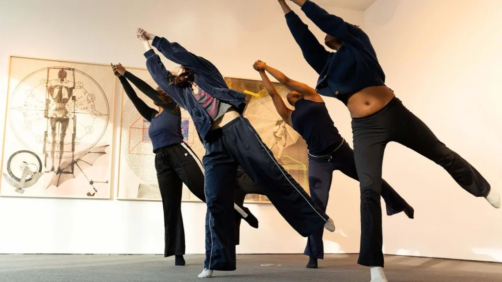 Four people in casual, dark clothing perform a contemporary dance pose indoors, each balancing on one leg with arms raised. Two large, abstract art pieces hang on the white wall behind them.