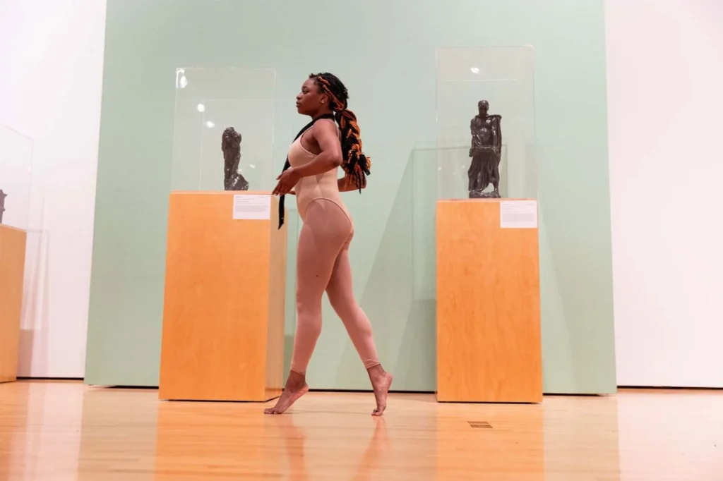 A ballet dancer in nude-colored attire and pointe shoes performs in a gallery space, standing on tiptoe between two wooden pedestals displaying sculptures in glass cases.