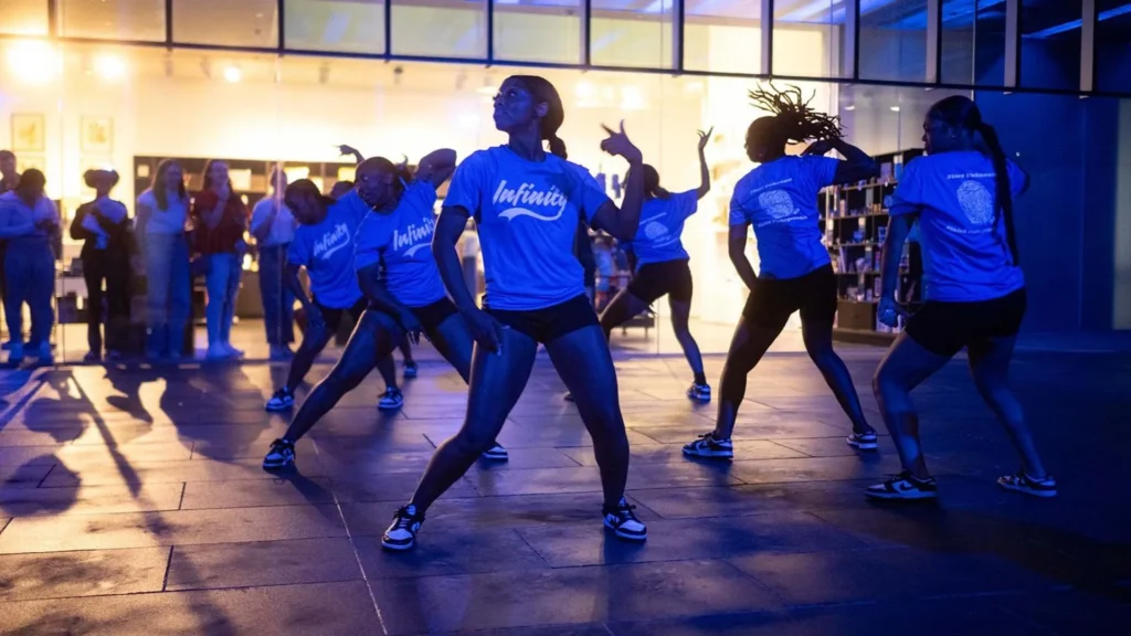 A group of dancers wearing matching blue Infinity shirts perform an energetic routine at night, illuminated by blue and yellow lights, while a crowd watches in the background.