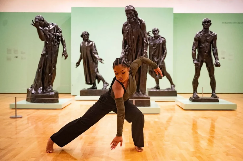 A dancer with braided hair and wearing dark clothing performs a graceful pose on a polished museum floor, with bronze statues arranged behind her against green walls.