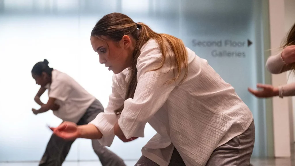 Three people perform a contemporary dance routine indoors, wearing white tops and gray pants. The background shows a sign for Second Floor Galleries on a glass wall.