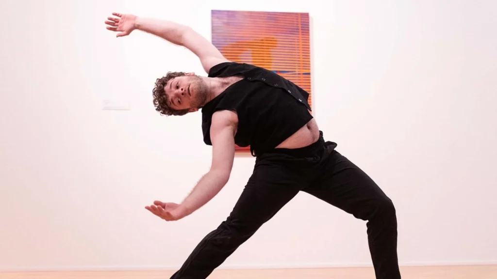 A person wearing black clothing performs a dynamic dance pose, leaning sideways with arms extended, in front of a colorful abstract artwork on a white gallery wall.
