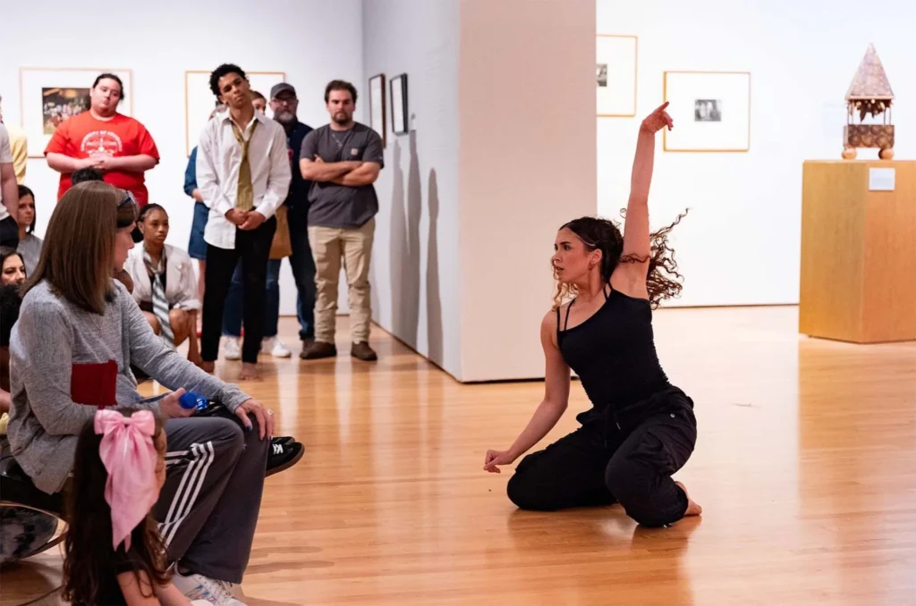 A woman in black clothes performs a dance on a museum floor, one arm raised, as an audience watches attentively. Framed art and sculptures are displayed on the walls and pedestals in the background.