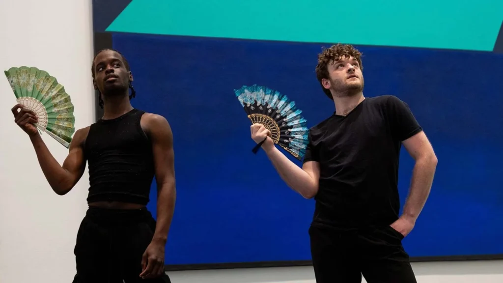 Two people in black outfits hold decorative fans and pose dramatically in front of a blue and teal geometric background.