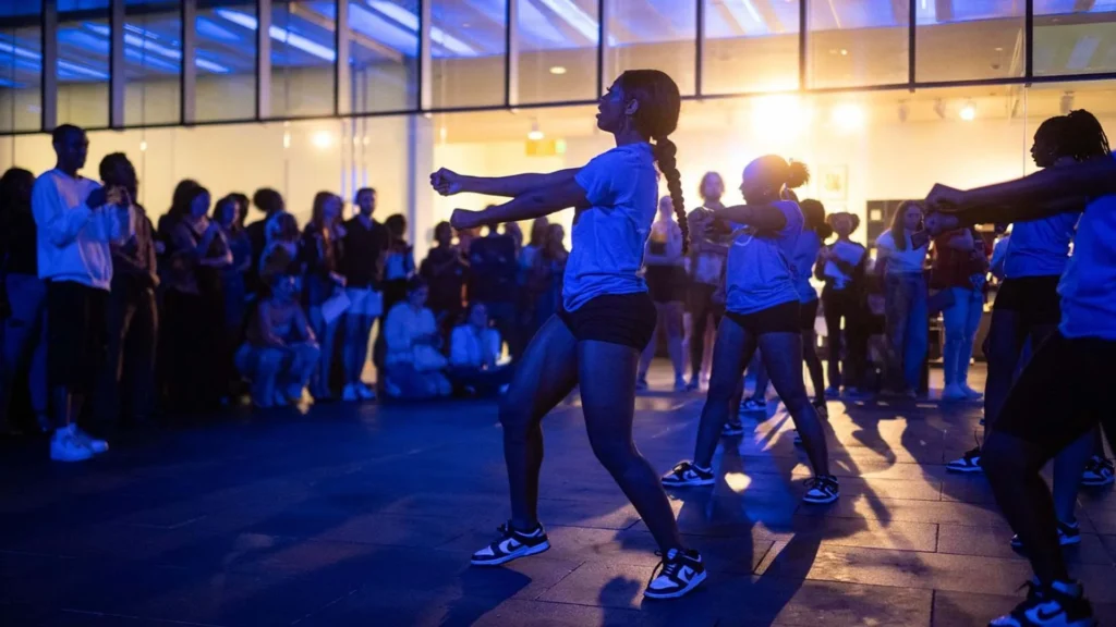 A group of young dancers performs energetically in matching outfits before a crowd indoors, with dramatic lighting casting blue and yellow tones across the scene.