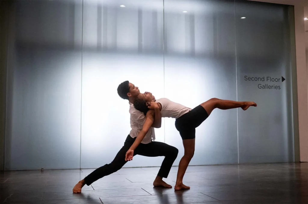 Two contemporary dancers perform an expressive duet indoors; one leans forward supporting the other, who extends one leg gracefully behind her. A sign reads Second Floor Galleries in the background.
