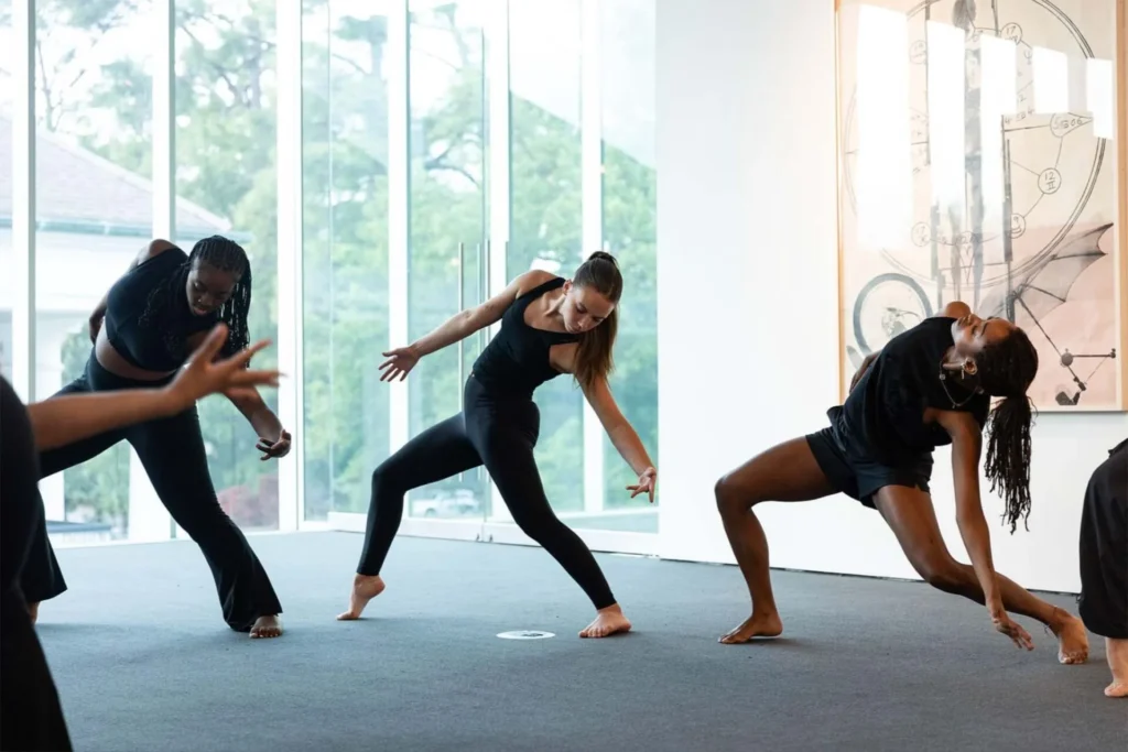 Three women in black dancewear perform expressive, contemporary dance moves in a bright room with large windows and modern art on the wall. Their poses suggest dynamic movement and balance.