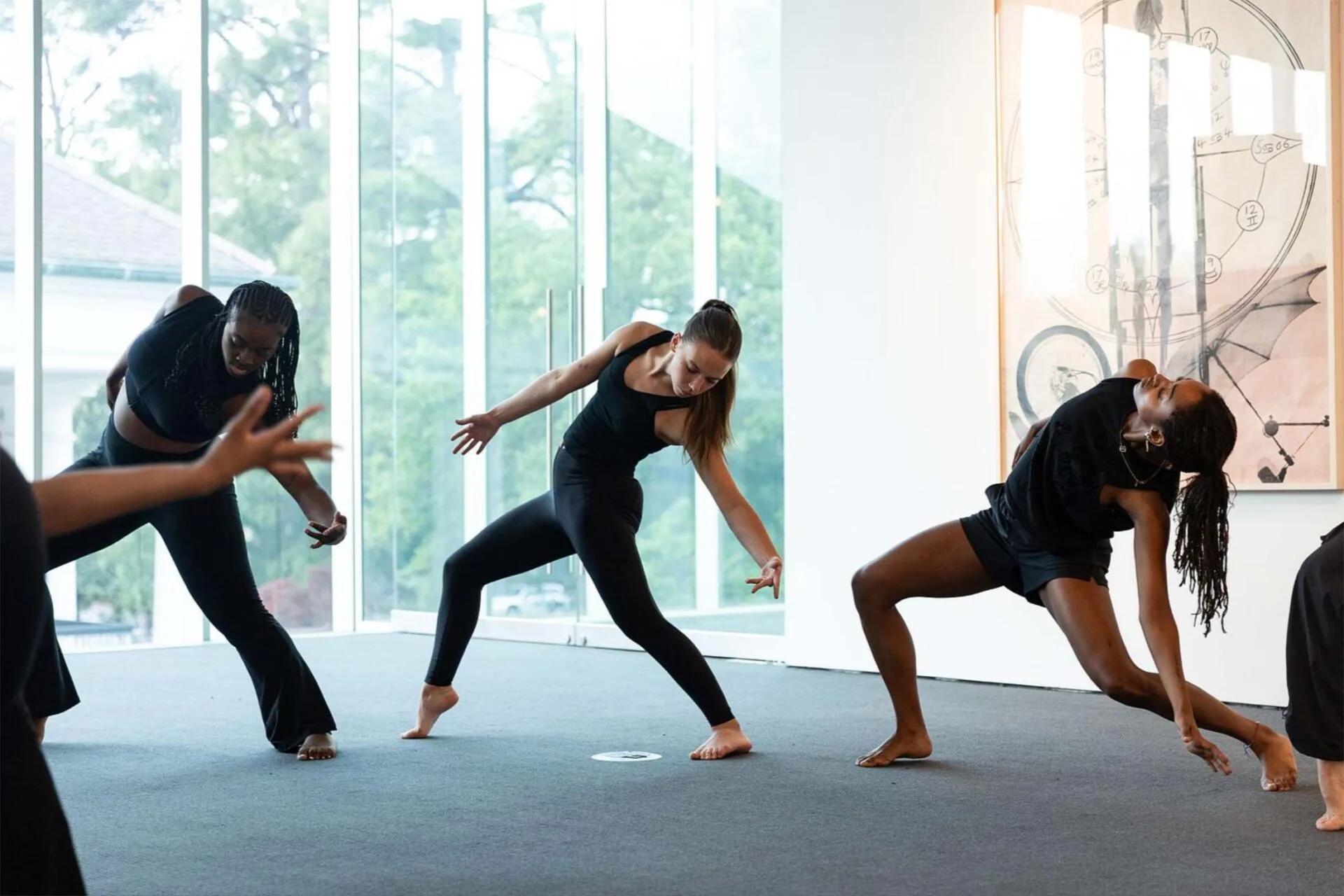 Three women in black dancewear perform expressive, contemporary dance moves in a bright room with large windows and modern art on the wall. Their poses suggest dynamic movement and balance.
