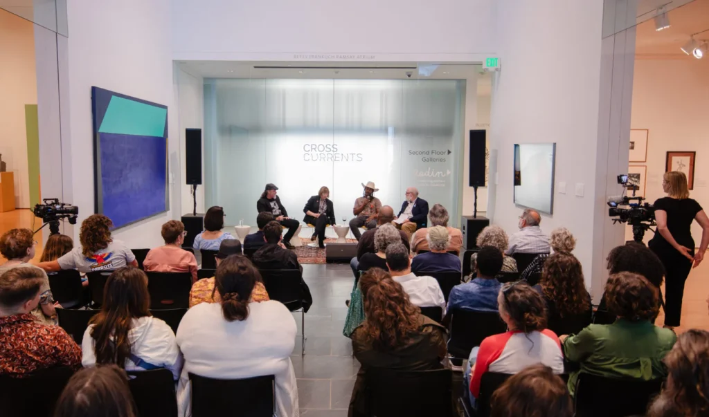 An audience watches a panel discussion in an art gallery. Five people sit at the front, speaking, with CROSS CURRENTS displayed on a glass wall behind them. Cameras and artwork are visible around the room.