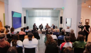 An audience watches a panel discussion in an art gallery. Five people sit at the front, speaking, with CROSS CURRENTS displayed on a glass wall behind them. Cameras and artwork are visible around the room.