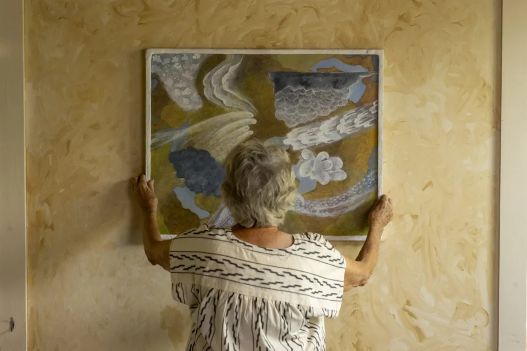 An older woman with gray hair, seen from behind, hangs a colorful abstract painting on a textured beige wall, wearing a light blouse with black stripes.