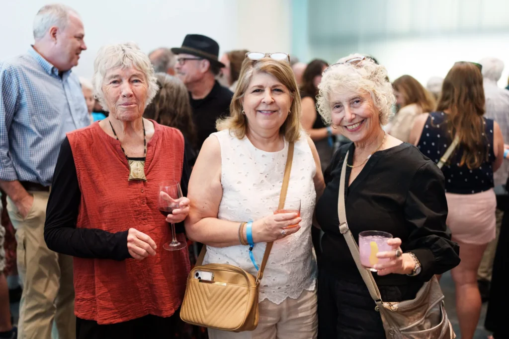Three smiling women stand close together at a social event, each holding a drink. Other people mingle in the background. The atmosphere appears lively and friendly.