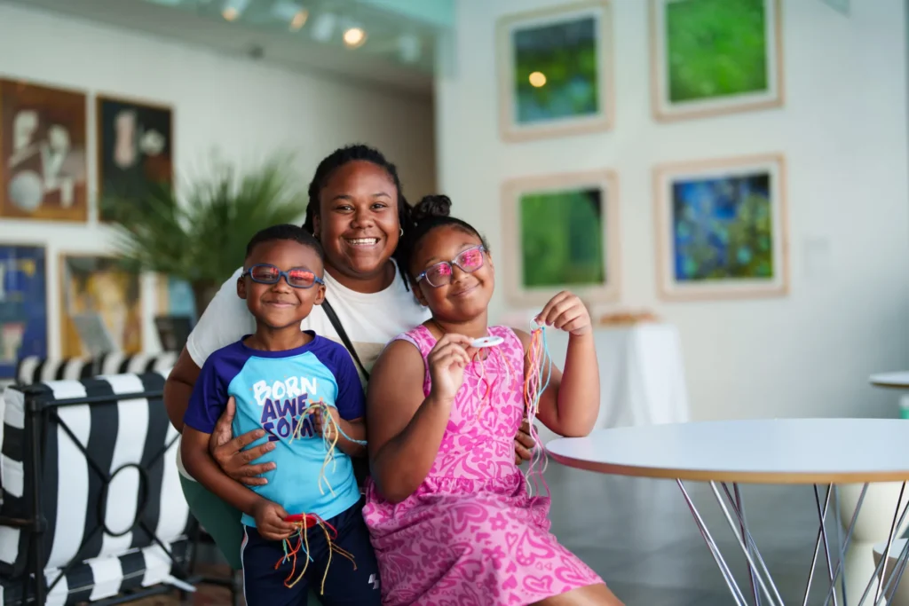 A smiling woman poses with two children indoors; the boy wears glasses and a blue shirt, the girl has glasses and a pink dress, both holding colorful string crafts. Artworks hang on the walls in the background.