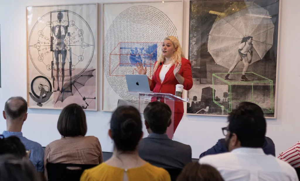 A woman in a red suit speaks at a clear podium to a seated audience in a gallery, with three large abstract art pieces hanging on the wall behind her.