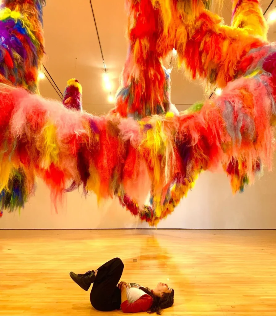 A person lies on a wooden floor in a gallery, looking up at large, colorful, fluffy yarn sculptures hanging from the ceiling, featuring vibrant reds, yellows, oranges, and purples.