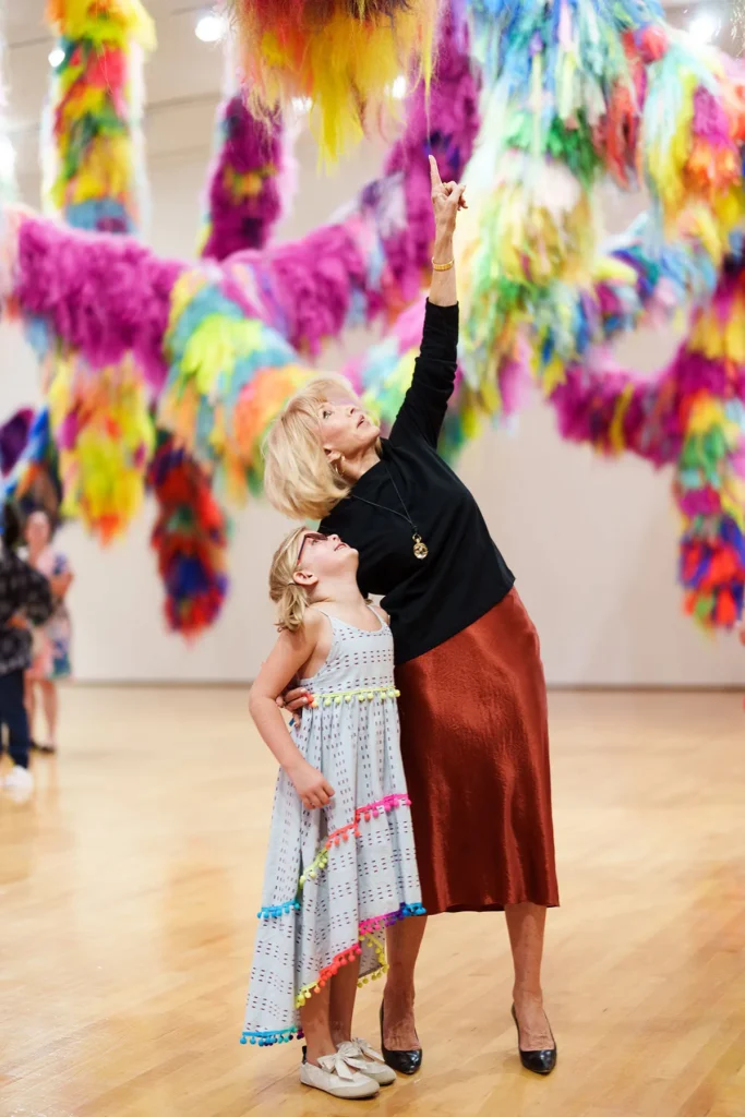 An older woman and a young girl stand in an art gallery, looking up. The woman points toward colorful, fluffy, hanging art installations above them. The girl gazes up, smiling, surrounded by vibrant decorations.