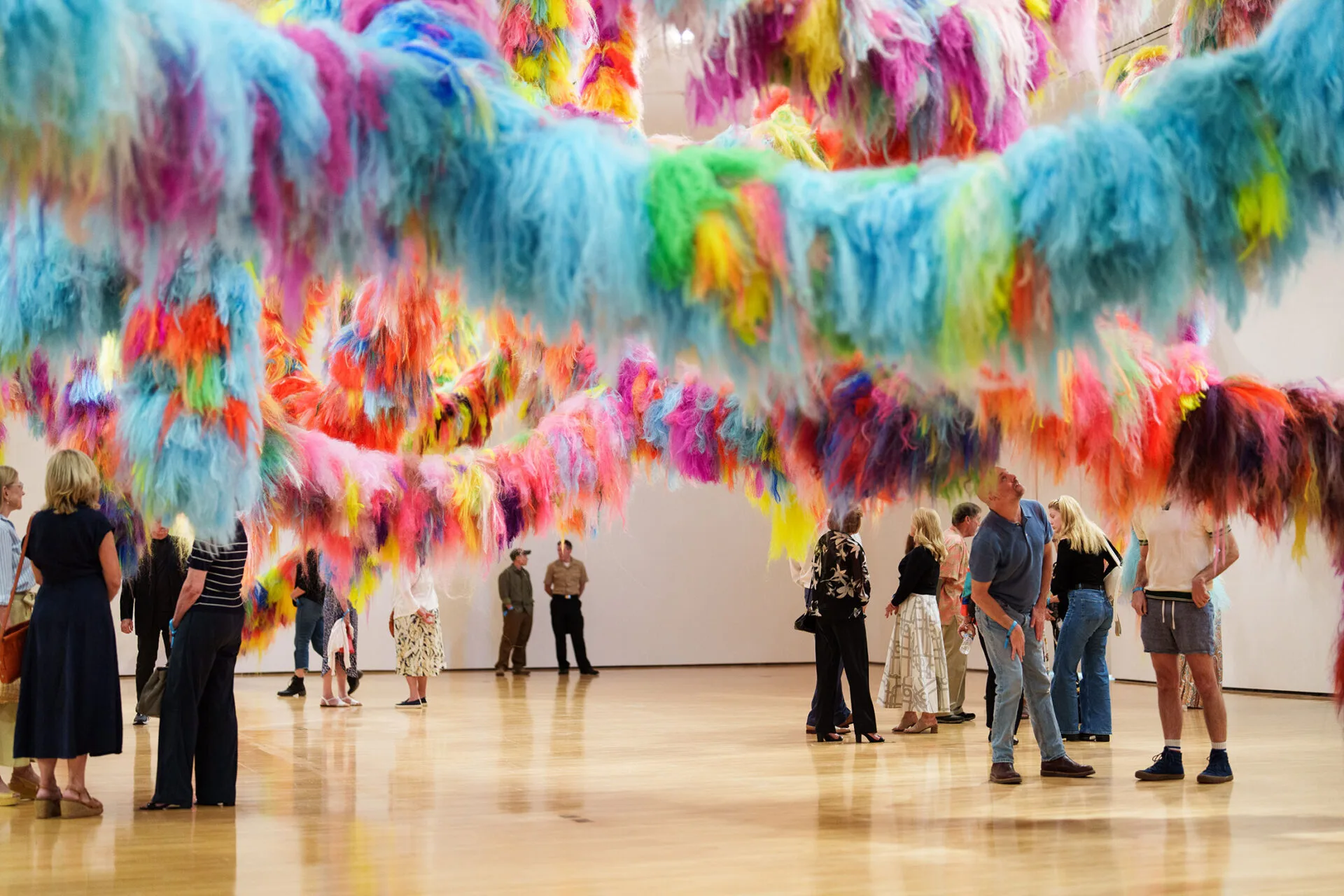 People explore an art gallery with large, colorful, fluffy, hanging installations in shades of pink, blue, yellow, green, and orange suspended from the ceiling above a polished wooden floor.