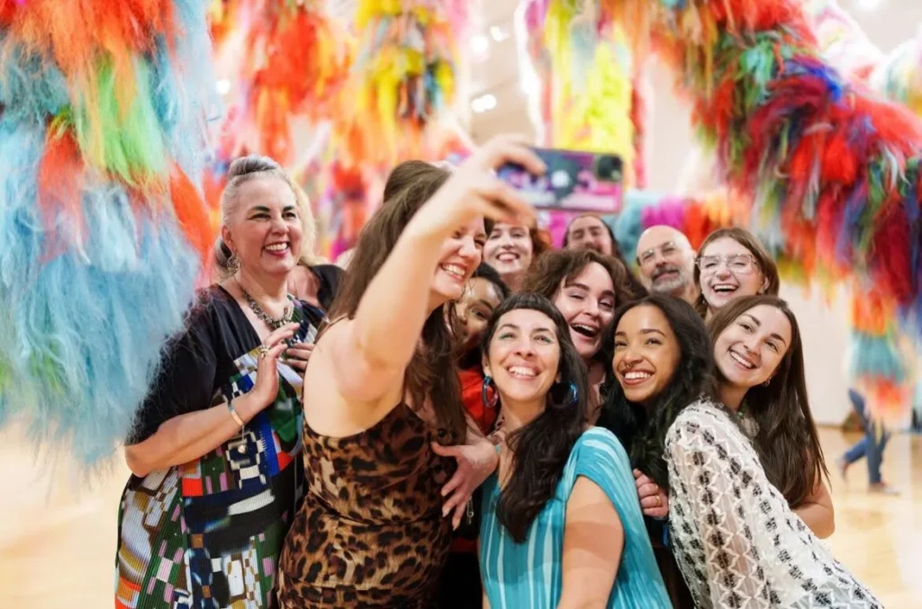 A group of people smile and pose for a selfie together in a colorful room decorated with hanging, furry, rainbow-colored art installations.
