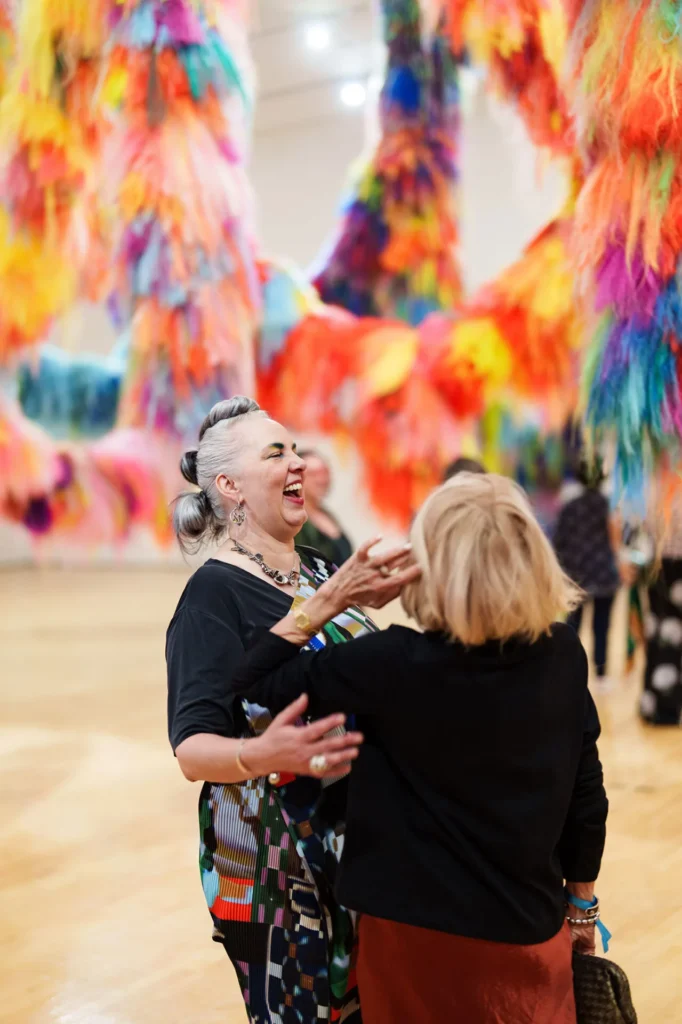 Two women laugh and embrace in a bright gallery filled with large, colorful, fuzzy art installations hanging from the ceiling. The atmosphere is lively and cheerful.