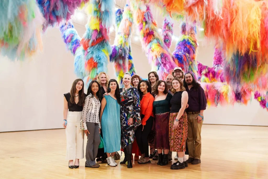 A group of people stand and smile together in a brightly lit gallery with large, colorful, fluffy hanging art installations in the background.