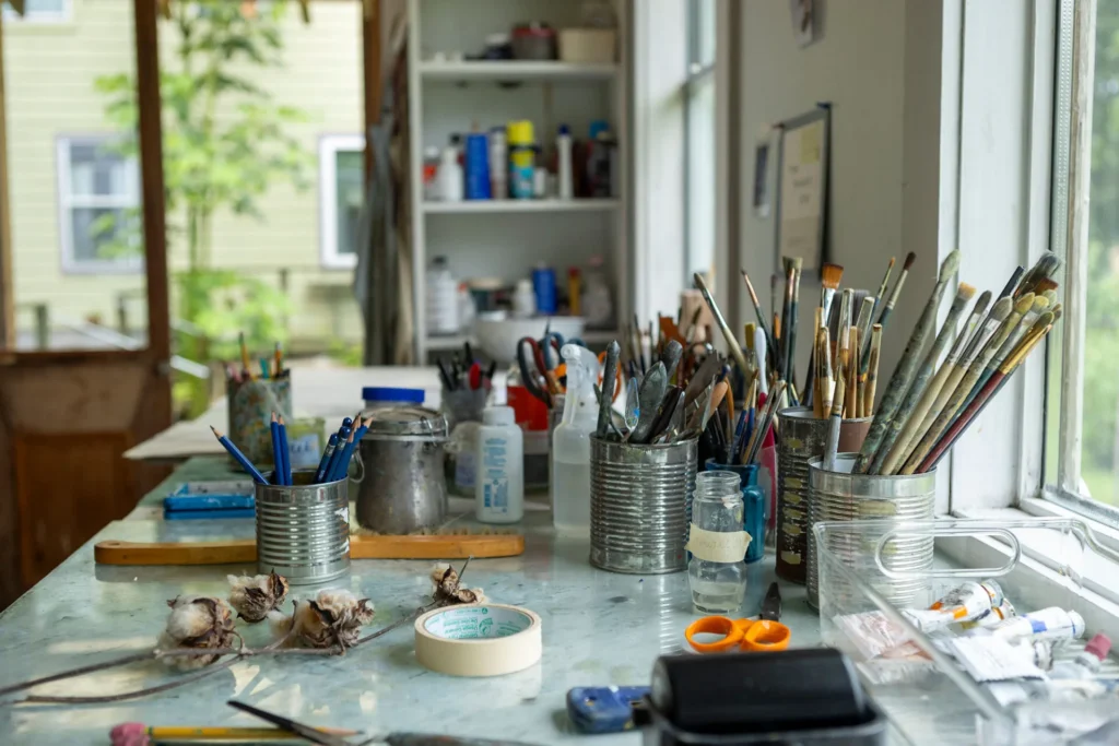 A cluttered art studio table with jars of paintbrushes, scissors, tape, and various art supplies sits by a window. Shelves with bottles and containers are visible in the background.