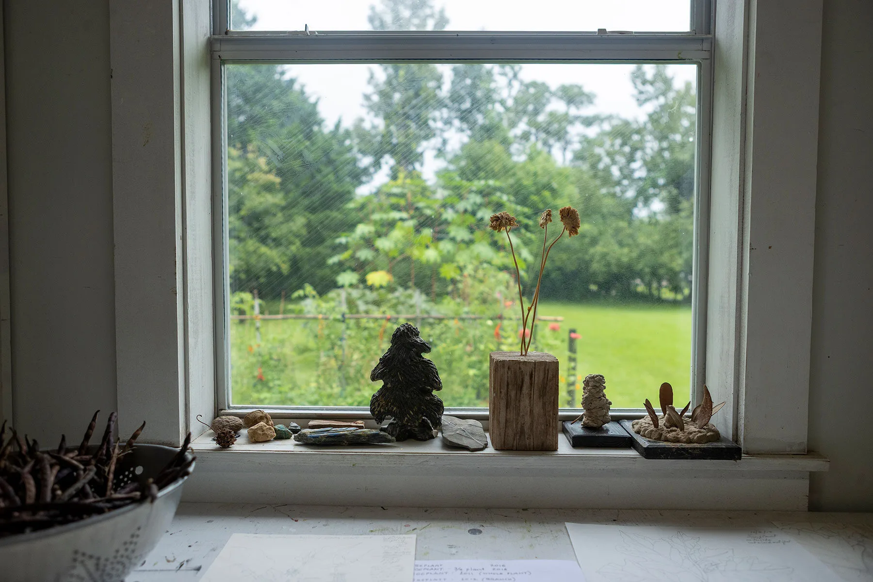 A windowsill displays small sculptures, rocks, twigs, and a wooden vase with dried flowers. Outside, a green garden and trees are visible through the window on a cloudy day. Papers and a bowl of sticks sit on the table below.