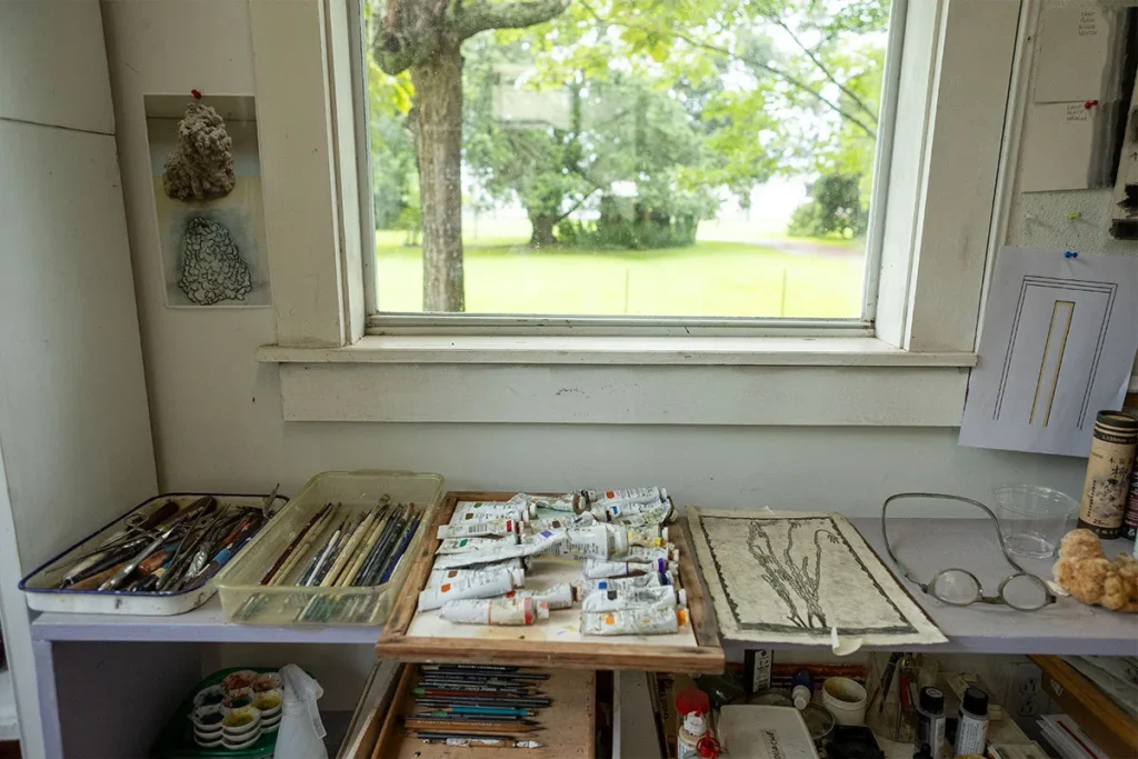 An artist’s workspace with paint tubes, brushes, and sketches on a table beneath a window, overlooking a green, tree-lined yard outside.